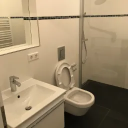 Biała umywalka z szafką, toaleta z otwartą klapą i szklana kabina prysznicowa na tle białych płytek z czarnym paskiem dekoracyjnym, czarne płytki podłogowe.