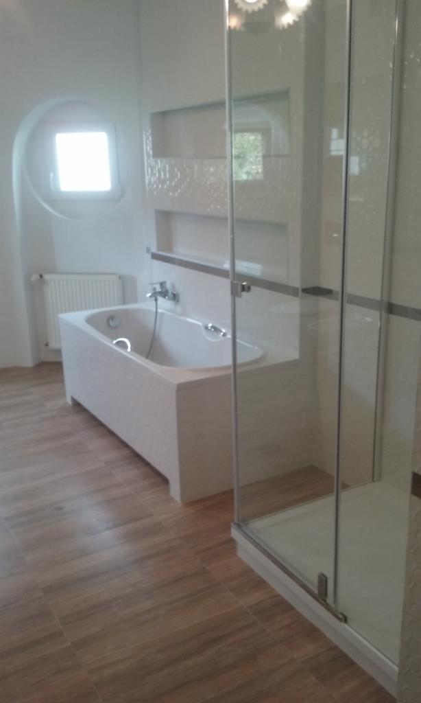 Nowoczesna łazienka z białą wanną obudowaną mozaiką, kabiną prysznicową ze szklanymi drzwiami i drewnopodobną podłogą w odcieniach brązu, oświetlona światłem wpadającym przez okrągłe okno.