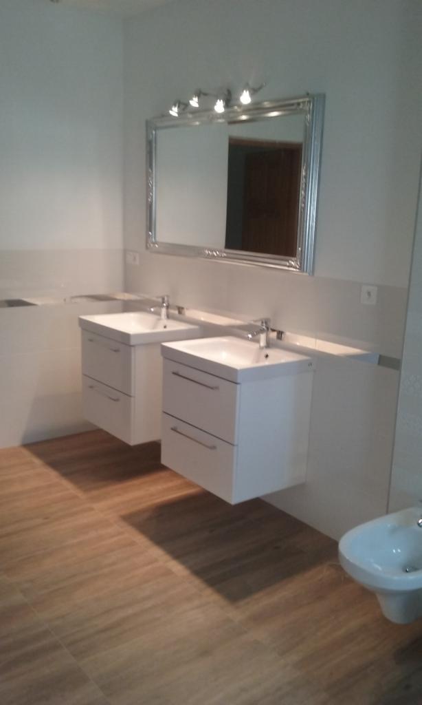 Nowoczesna łazienka z dwoma białymi szafkami pod umywalki, lustrem w srebrnej ramie i bidetem na podłodze z paneli imitujących drewno.