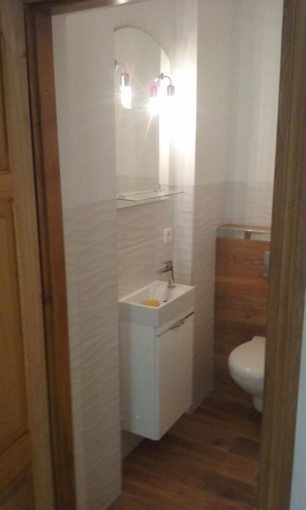 Mała łazienka z umywalką nablatową na szafce, owalnym lustrem z podwójnym oświetleniem i toaletą z drewnianą obudową spłuczki, wykończona płytkami z delikatnym, falistym wzorem.