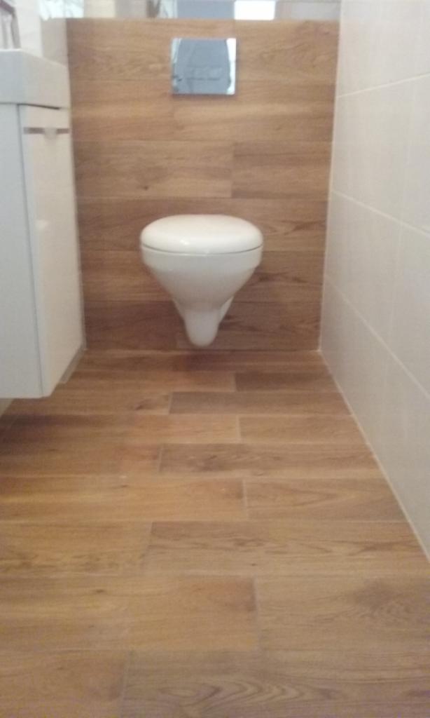 Nowoczesna toaleta wisząca zamontowana na ścianie wyłożonej płytkami imitującymi drewno, z podłogą wyłożoną identycznymi płytkami, obok znajduje się biała szafka z umywalką i białe płytki na ścianie.