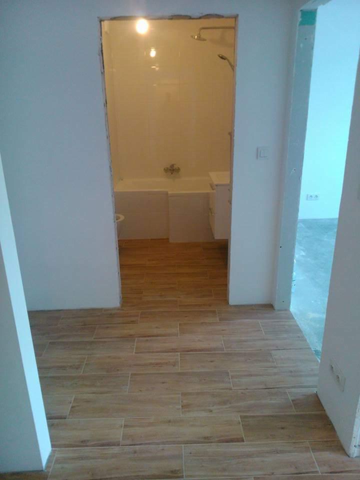 Ułożona podłoga z płytek imitujących drewno prowadząca do łazienki z wanną i umywalką, widok przez framugę drzwi, ściany w kolorze białym i jasnoniebieskim.