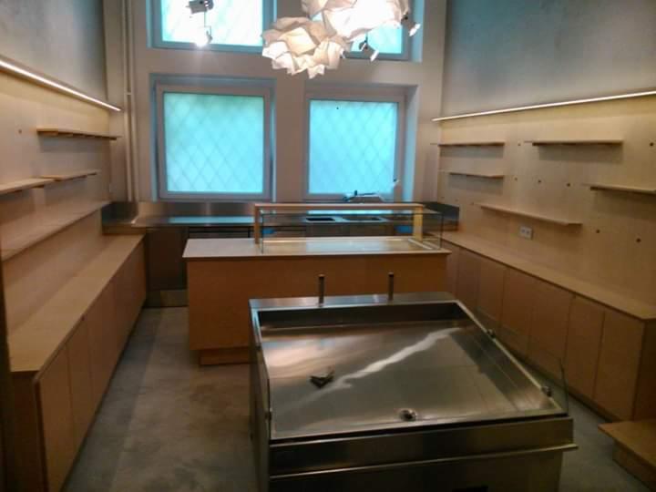 Surowe wnętrze sklepu spożywczego w trakcie wykańczania: drewniane regały i szafki wzdłuż ścian, stalowe lady chłodnicze, oświetlenie LED, sufit z nowoczesną lampą.