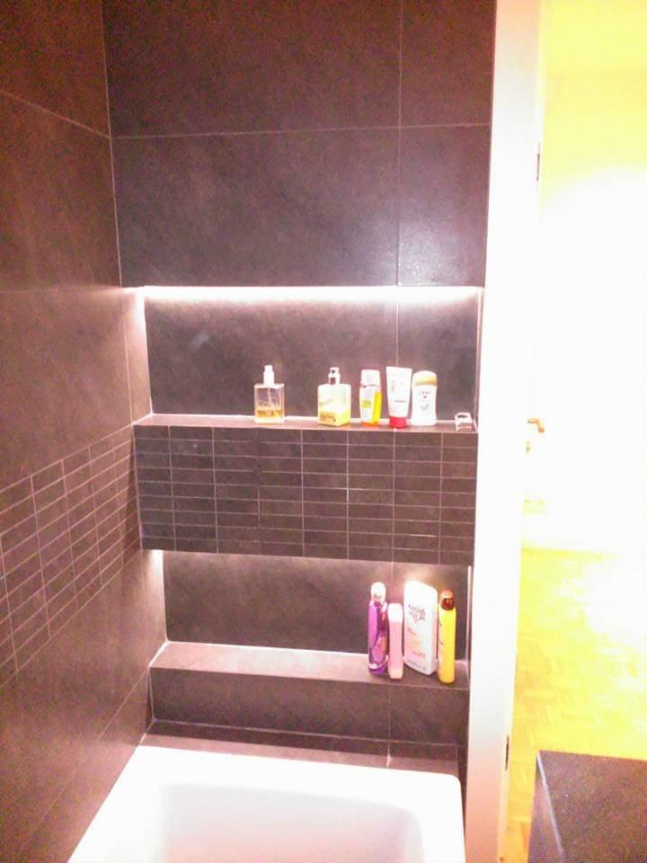 Łazienka z ciemnymi płytkami, podświetlanymi półkami wnękowymi z kosmetykami i białą wanną, widok z góry na część wanny.