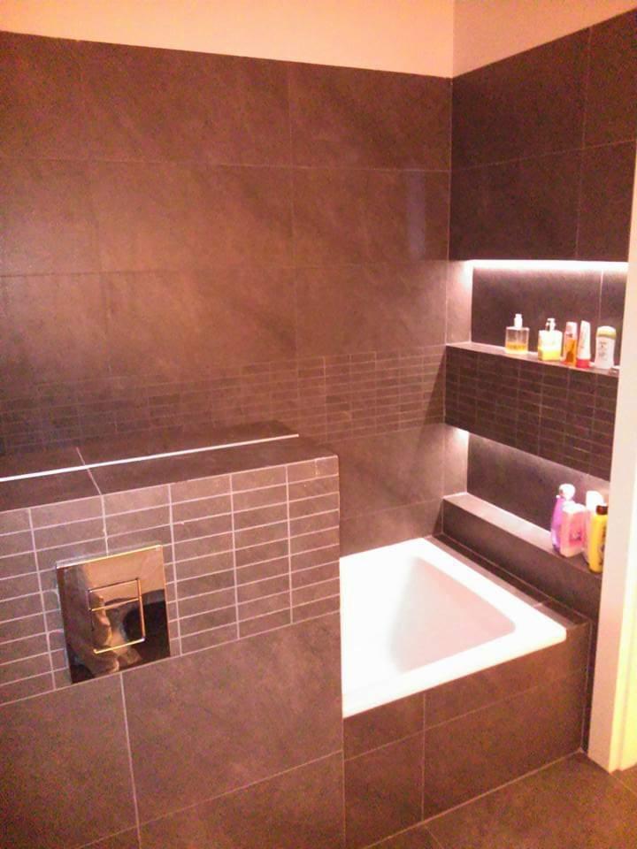 Nowoczesna łazienka z ciemnymi płytkami imitującymi kamień, podświetlane wnęki z kosmetykami i biała wanna.