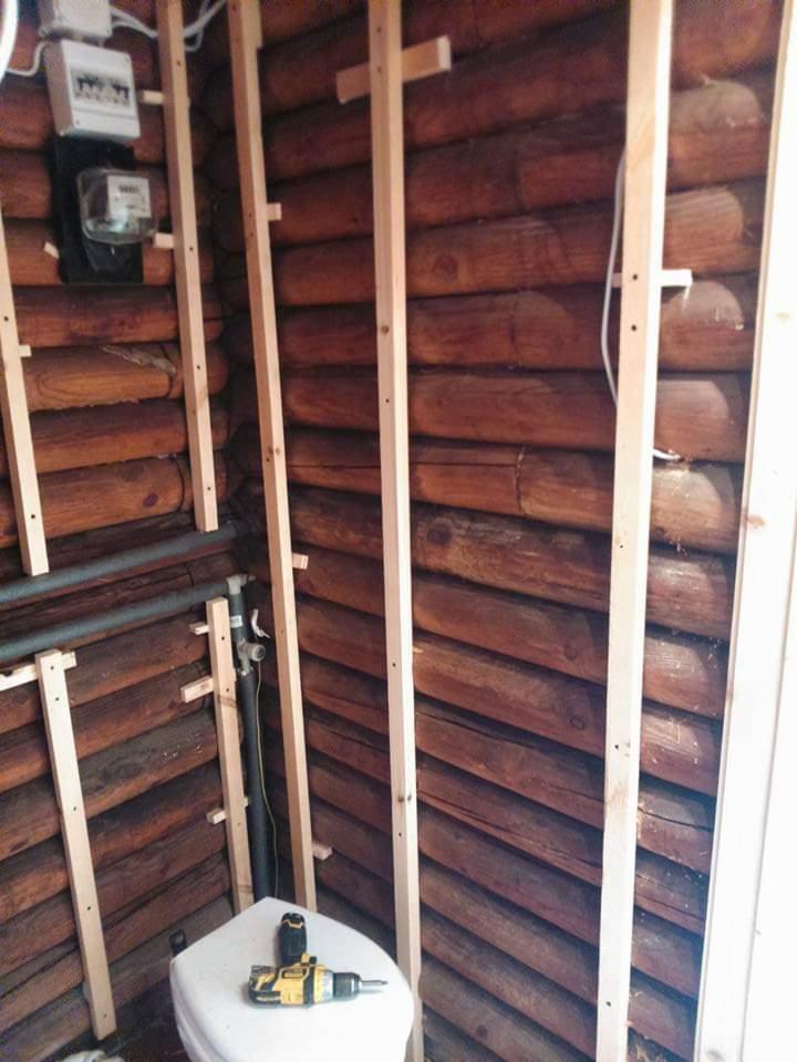 Wnętrze łazienki w trakcie prac, drewniane ściany z bali, pionowe listwy montażowe, biała umywalka z wiertarką akumulatorową, widoczne rury i licznik elektryczny.