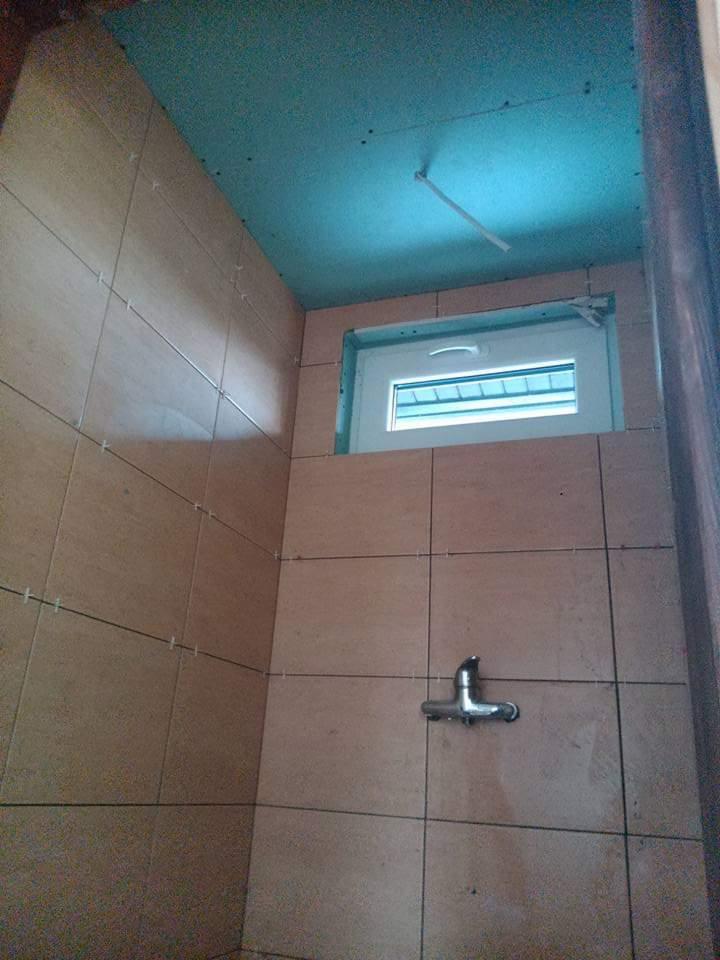 Ściana łazienki w trakcie kładzenia płytek, widoczny fragment okna, sufit z płyt gipsowo-kartonowych pomalowany na niebiesko i zamontowana bateria.