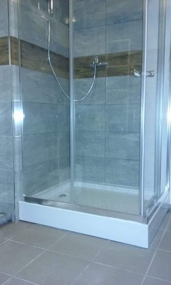 Szklana kabina prysznicowa z brodzikiem, wykończona szarymi płytkami z drewnianym akcentem, zamontowana w łazience z szarą podłogą.