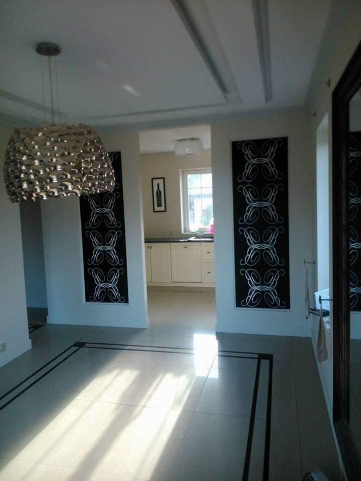 Jasne wnętrze z kremowymi płytkami podłogowymi z czarnymi obramowaniami, dekoracyjnymi panelami ściennymi z białym ornamentem i nowoczesną lampą wiszącą.