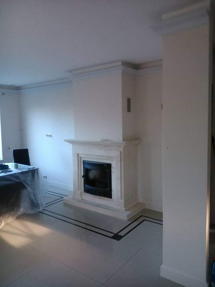 Nowoczesny kominek z czarnym wkładem, obudowany jasnym kamieniem, w minimalistycznym wnętrzu z listwami przypodłogowymi i dekoracyjnymi na suficie, wykończony jasnymi płytkami podłogowymi...