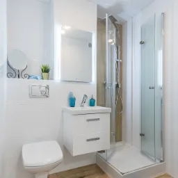 Jasna, nowoczesna łazienka z białymi płytkami, drewnopodobną podłogą, wiszącą szafką z umywalką, podświetlanym lustrem oraz kabiną prysznicową ze szklanymi drzwiami.