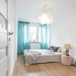 Vip Building - Jasna sypialnia z drewnianą podłogą, białymi ścianami, turkusowymi zasłonami, łóżkiem z narzutą, lampką nocną na różowym pufie i nowoczesnym oświetleniem sufitowym.