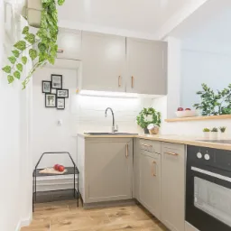 Jasna kuchnia w odcieniach szarości z drewnianymi akcentami: szafki z miedzianymi uchwytami, drewniany blat, płytki podłogowe imitujące drewno, czarny zlew i piekarnik, dekoracje roślinne i ramki...