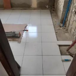 Białe płytki podłogowe w trakcie układania, widoczny przecinak do płytek na kartonie, nieotynkowane ściany z instalacją hydrauliczną w tle, widok zza framugi drzwi.