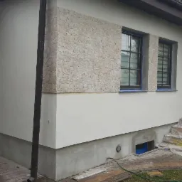 Fragment elewacji domu z oknami po remoncie, z częściowo wykończoną okładziną imitującą kamień, zabezpieczonymi taśmą okiennicami i widocznym fragmentem rynny.