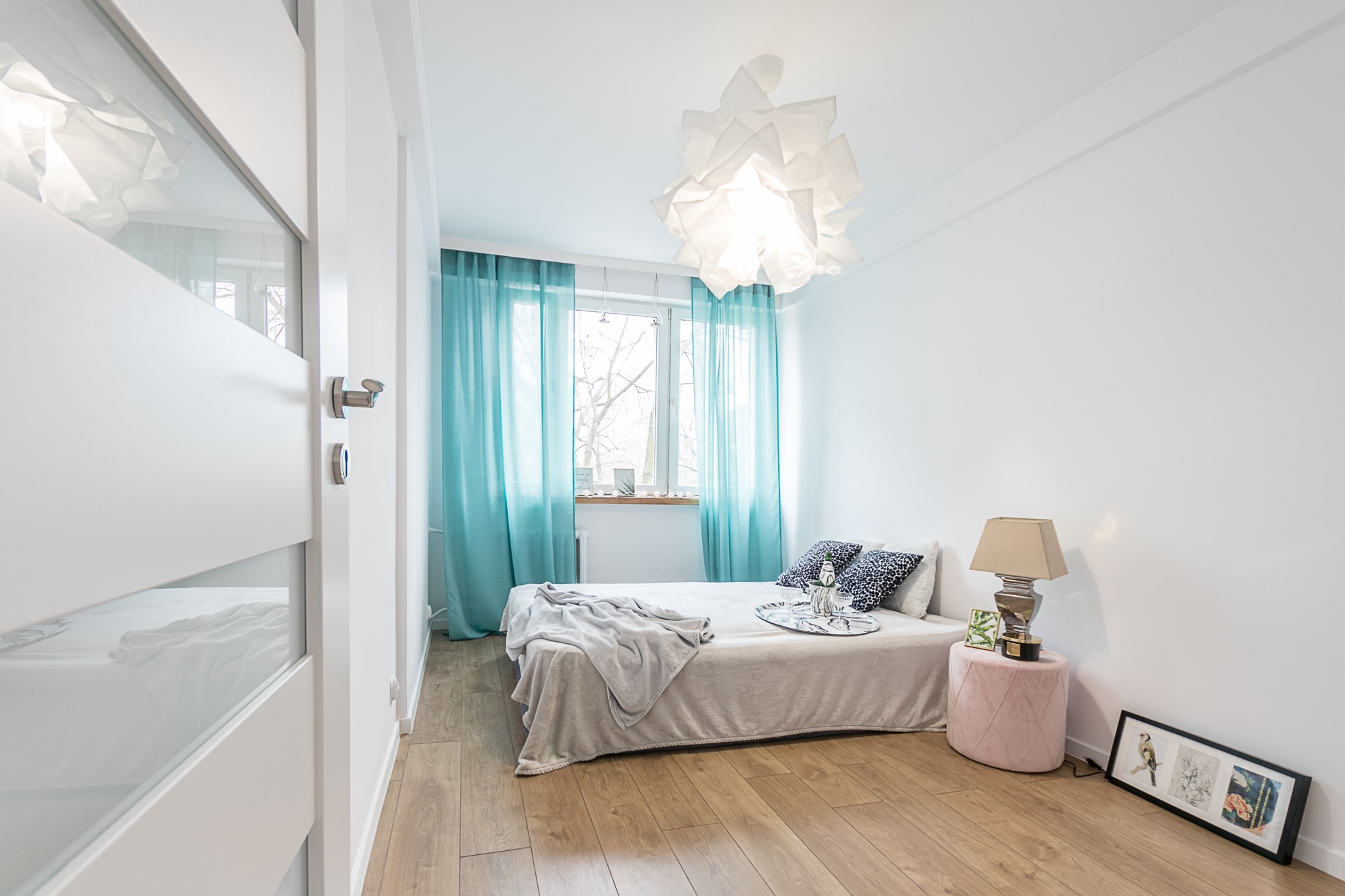 Jasna sypialnia z drewnianą podłogą, białymi ścianami, turkusowymi zasłonami, łóżkiem z narzutą, lampką nocną na różowym pufie i nowoczesnym oświetleniem sufitowym.