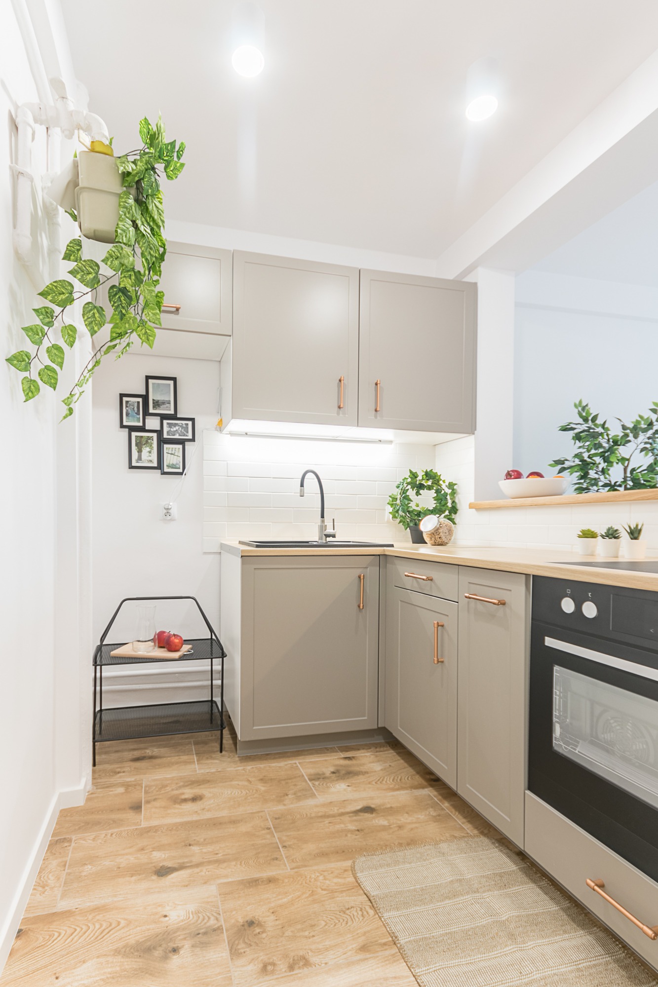 Jasna kuchnia w odcieniach szarości z drewnianymi akcentami: szafki z miedzianymi uchwytami, drewniany blat, płytki podłogowe imitujące drewno, czarny zlew i piekarnik, dekoracje roślinne i ramki...