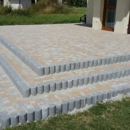 Trzy stopnie schodów prowadzące na taras z beżowo-brązowej kostki brukowej, obrzeża z szarych betonowych palisad, widok z poziomu trawnika.