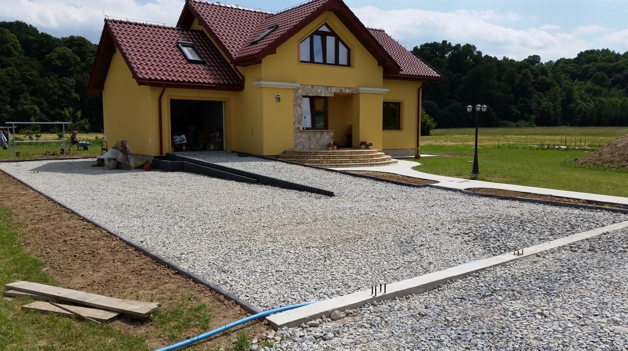 Żwirowy podjazd przed żółtym domem z czerwoną dachówką, widoczne obrzeża i przygotowane podłoże, fragment niebieskiego węża.