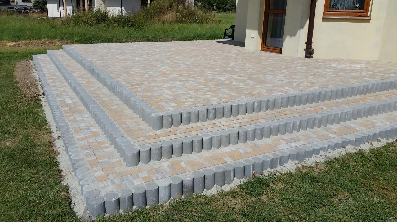 Trzy stopnie schodów prowadzące na taras z beżowo-brązowej kostki brukowej, obrzeża z szarych betonowych palisad, widok z poziomu trawnika.