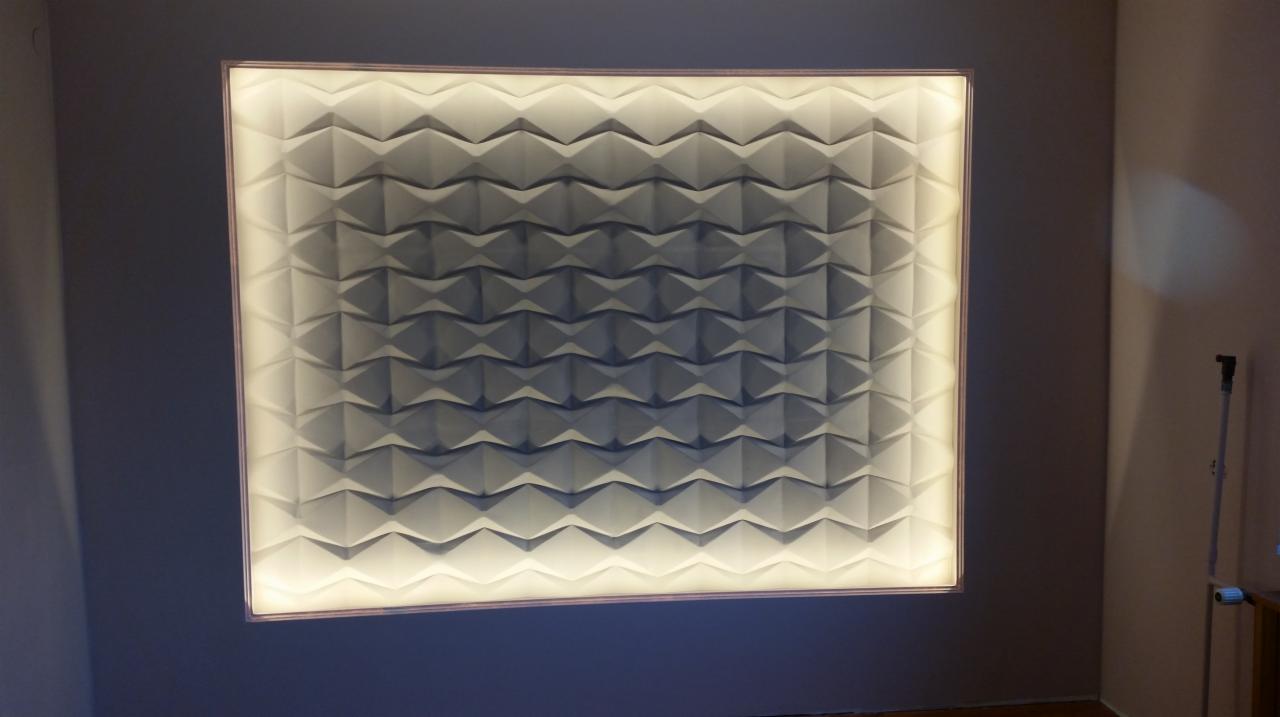 Panel ścienny 3D z podświetleniem LED, tworzący geometryczny wzór na szarej ścianie w pomieszczeniu.