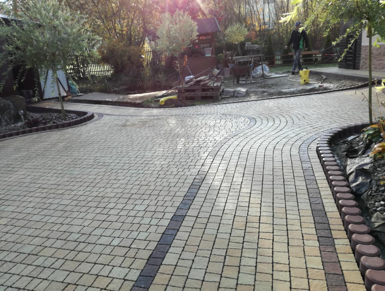 Ułożona kostka brukowa w jasnych odcieniach z ciemniejszymi pasami i obrzeżami, na posesji z drzewami i elementami małej architektury, widoczny mężczyzna w tle.