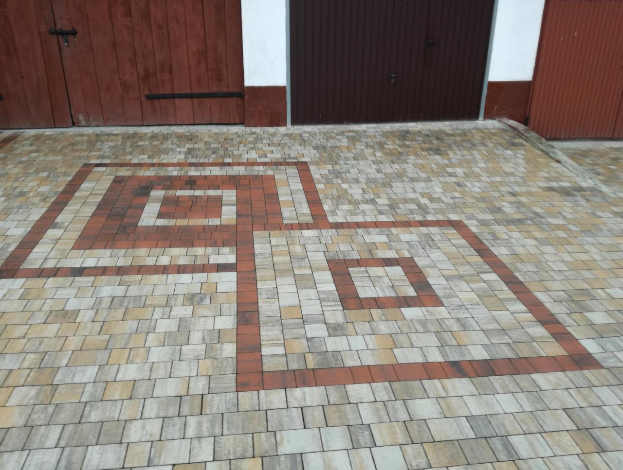 Układanka z kostki brukowej w odcieniach beżu i brązu, tworząca geometryczny wzór na podjeździe przed garażem z drewnianymi drzwiami.