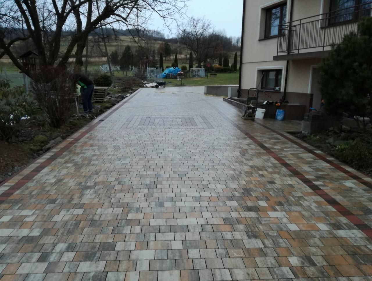 Nowo ułożona, wielobarwna kostka brukowa na podjeździe przed domem, z widocznym wzorem w centralnej części i bordiurą po bokach, w tle pracownik i sprzęt budowlany.