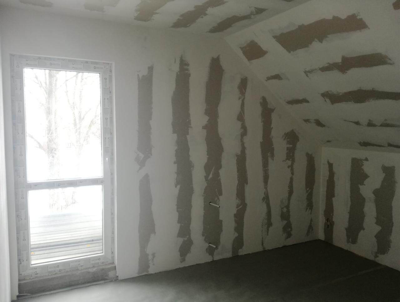 Pokój na poddaszu w trakcie prac wykończeniowych: ściany i skośny sufit z nałożoną warstwą gipsu przygotowane do dalszych prac, widoczne niedoskonałości i szpachlowane pasy, okno z widokiem na drzewa.