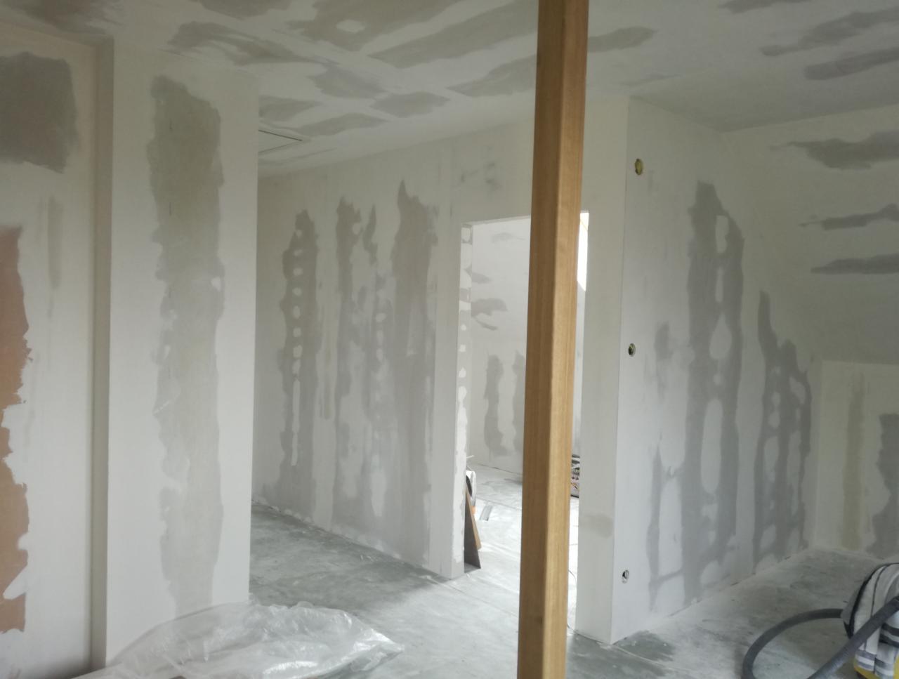 Wnętrze w trakcie remontu, ściany i sufit pokryte świeżo nałożoną warstwą gładzi szpachlowej, widoczny drewniany element konstrukcyjny i sprzęt remontowy.