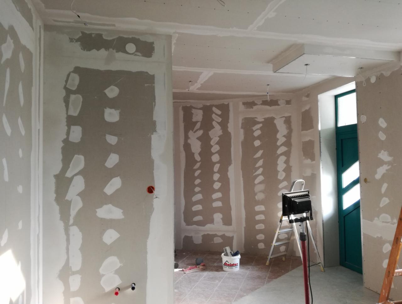 Wnętrze w trakcie remontu: ściany pokryte płytami gipsowo-kartonowymi ze śladami szpachlowania, widoczna drabina, wiadro z masą szpachlową i lampa budowlana.