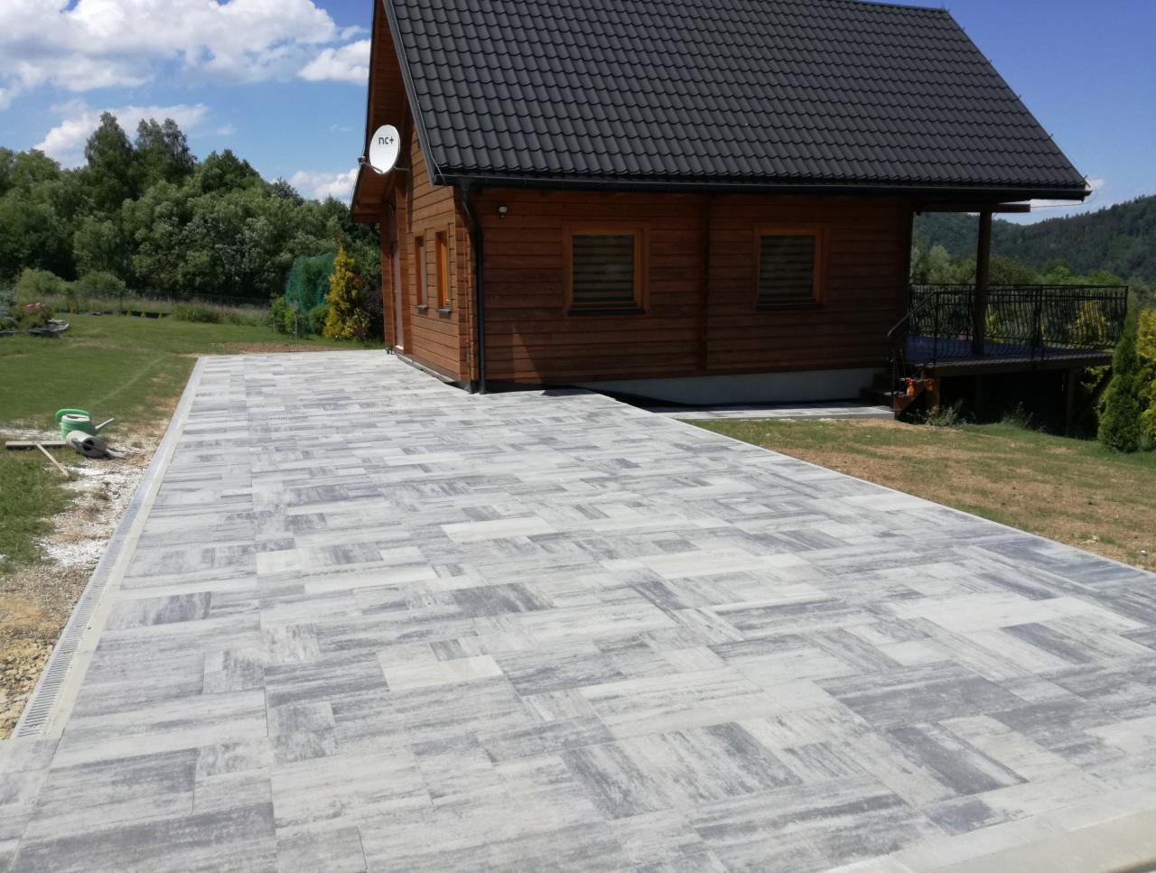 Ułożona szara kostka brukowa prowadząca do drewnianego domu z czarnym dachem, widok z poziomu gruntu, w tle zieleń drzew i wzgórza.