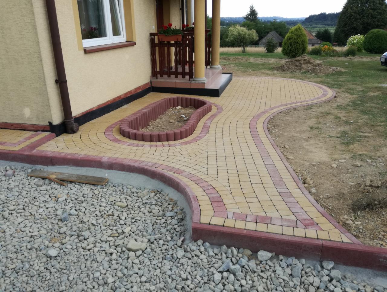 Żółto-czerwona kostka brukowa ułożona w zakrzywiony chodnik prowadzący do wejścia domu z kolumnami, z elementami dekoracyjnymi i przygotowanym miejscem na rabatę, częściowo podsypana kamieniami.