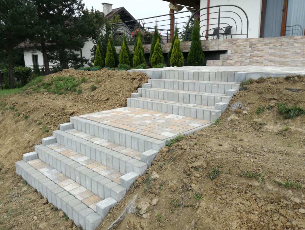 Schody zewnętrzne z kostki brukowej prowadzące do tarasu domu, widoczne ślady prac ziemnych.