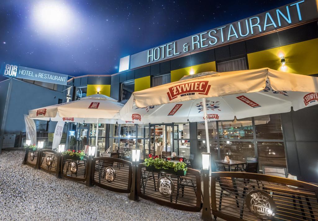 Oświetlony ogródek restauracyjny z parasolami Żywiec i drewnianymi barierkami z logo, widok na hotel nocą.