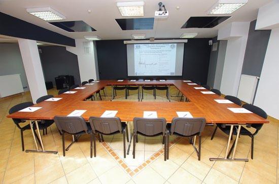 Sala konferencyjna z układem stołów w kształcie litery U, rzutnik wyświetlający prezentację na ekranie, przygotowane miejsca z kartkami i długopisami.