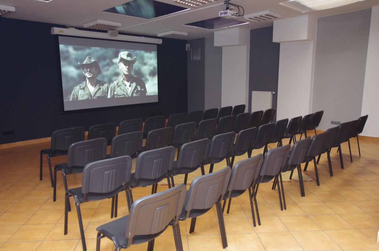 Sala konferencyjna z rzędami krzeseł ustawionych przed ekranem projekcyjnym wyświetlającym film z dwoma mężczyznami w mundurach i kapeluszach.