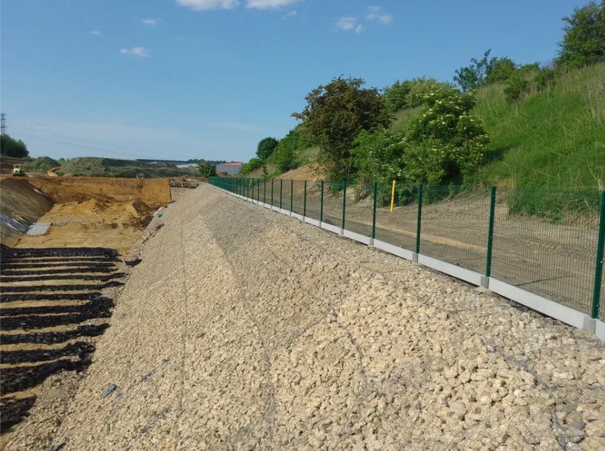 Zielone ogrodzenie panelowe z podmurówką betonową, zabezpieczające skarpę wysypaną kamieniami, w tle wykop i zieleń.