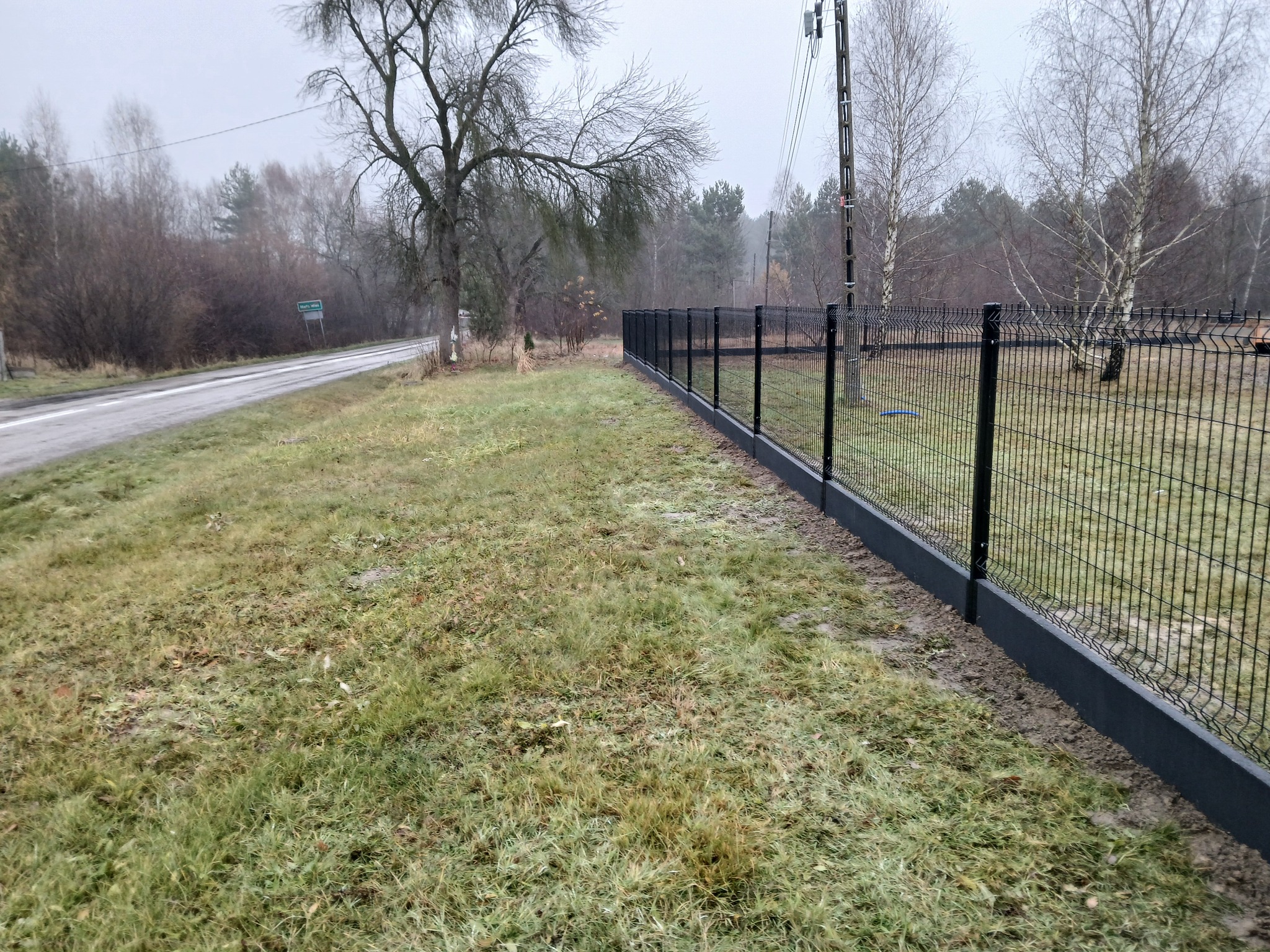 Czarny panelowy płot z betonową podmurówką, widok z boku na tle jesiennego krajobrazu i drogi.