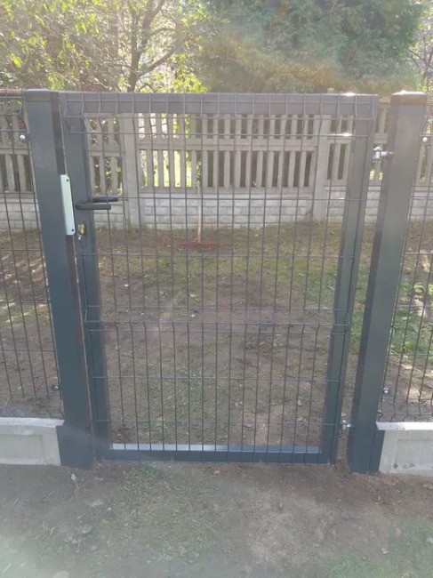 Szara, metalowa furtka panelowa z zamkiem i klamką, osadzona w betonowej podmurówce, widoczna na tle betonowego ogrodzenia i zieleni.
