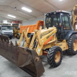 Żółto-szara koparko-ładowarka Case 580 Super LE 4x4 z dużym, ząbkowanym łyżką z przodu, zaparkowana w hali z innymi maszynami budowlanymi i ciężarówkami w tle.