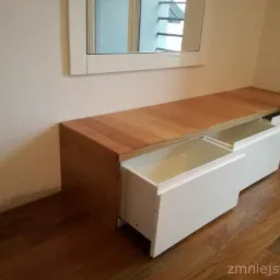 Niska komoda z drewna i białego lakieru z dwiema wysuniętymi szufladami, widoczna na tle drewnianej podłogi i ściany z lustrem.