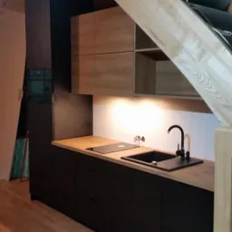 Nowoczesna kuchnia w aneksie pod schodami: czarne szafki dolne i drewnopodobne górne, drewniany blat, czarny zlew i bateria, oświetlenie punktowe pod szafkami, widoczne drewniane schody.