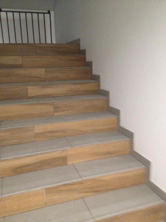 Schody wykończone płytkami imitującymi drewno, szare płytki na stopniach, czarna metalowa balustrada w tle, jasne ściany.