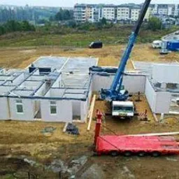 Montaż prefabrykowanego domu modułowego na przygotowanym terenie, z użyciem niebieskiego dźwigu i czerwonej platformy transportowej, widok z góry na plac budowy.