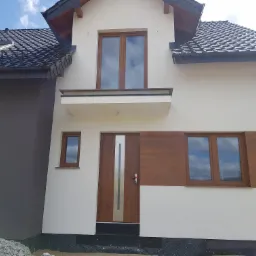 Nowoczesny dom jednorodzinny z jasną elewacją, brązowymi oknami i drzwiami, oraz ciemnoszarą częścią garażową. Widoczne prace wykończeniowe wokół budynku.