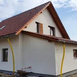 Nowo wybudowany dom z czerwonym dachem, jasną elewacją i żółtymi rurami spustowymi, widok z boku na tle błękitnego nieba.