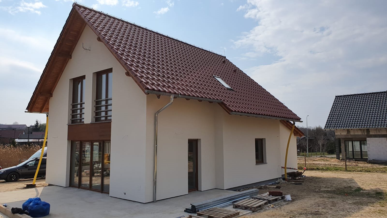 Nowoczesny dom murowany z brązową dachówką i jasną elewacją, widok z zewnątrz, z elementami wykończeniowymi w trakcie realizacji, w tle drugi budynek w stanie surowym.