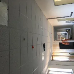 Korytarz w trakcie montażu sufitu podwieszanego z płyt o fakturze drobnego ziarna, widoczne niedokończone instalacje i przewody elektryczne. Część paneli zabezpieczona folią ochronną.