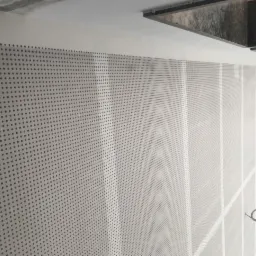 Ściana pokryta perforowaną płytą w trakcie montażu, widoczne okna i fragment sufitu, przewody elektryczne zwisające ze ściany.